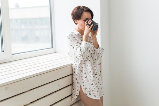 Beautiful young woman with cup of tea standing near the window at home