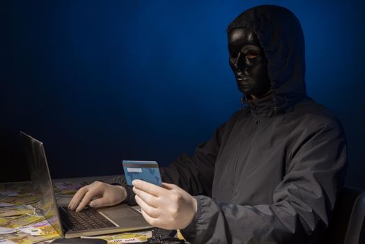Anonymous hacker in mask programmer uses a laptop to hack the system in the dark. The concept of cybercrime theft of money from bank cards