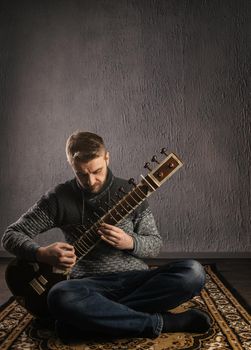 Portrait of a European man playing the sitar sitting on the carpet- Image