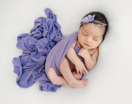 Adorable newborn baby girl sleeping swaddled in fabric. Cute infant kid child napping with wreath on her head during studio photoshoot