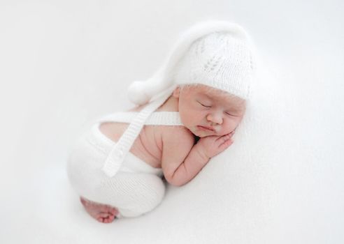 Newborn baby boy sleeping wearing knitted white costume and hat. Infant child kid napping during studio photoshoot