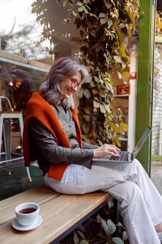 Happy mature woman with glasses and cup of coffee types on laptop keyboard sitting on bench near cafe window on autumn day