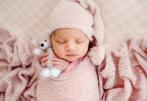 Adorable newborn baby girl sleeping wearing hat and holding knitted teddy bear toy. Cute infant kid child napping swaddled in fabric