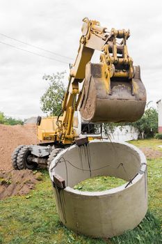 Hydraulic piston system excavator with a bucket, lifting on steel cable concrete sewer ring. Repairs or sewage works on an industrial site.