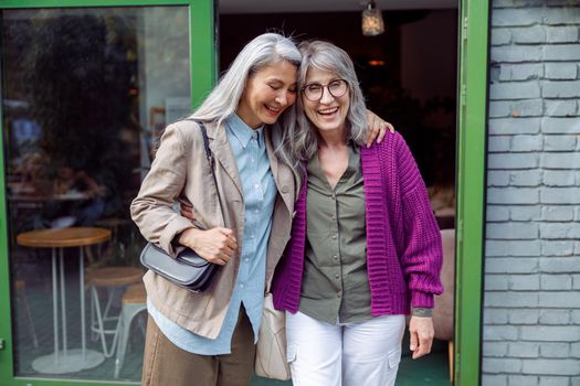 Pretty senior Asian lady hugs grey haired friend in purple jacket standing near cafe entrence on city street. Long-time friendship relationship