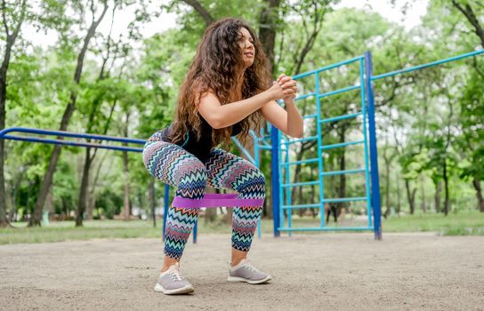 Girl with elastic rubber band doing squats exercising outdoors. Young woman training with sport equipment