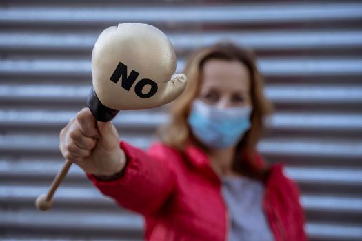 Young woman in protective sterile medical mask on her face, on striped background, holding small boxing glove with No sign. America president election, pandemic coronavirus concept.