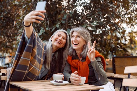 Joyful senior women friends take selfie with modern smartphone sitting at small table in street cafe on nice autumn day