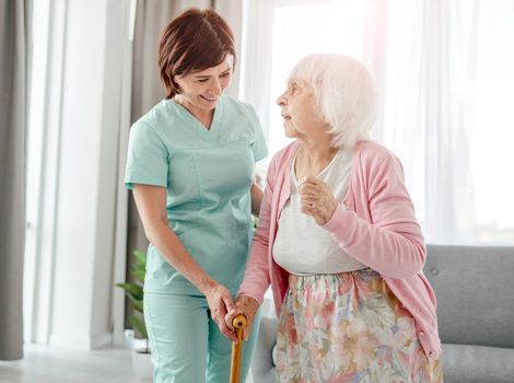 Young nurse helps elderly woman at home. Healthcare worker girl cares about senior female person indoors