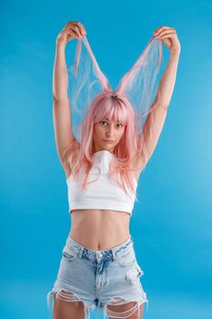 Funny female model in casual wear playing with pink hair, holding strands of it up over her head, standing isolated over blue studio background. Beauty, hair care concept