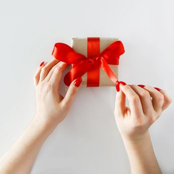 Gift with a red ribbon in hands on a white background. Gift in craft paper with a red ribbon in the hands of a young woman. Unpacking a holiday gift for Valentine's Day.