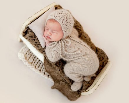 Newborn baby boy sleeping in tiny bed wearing knitted beige costume. Infant child kid napping on his tummy during studio photoshoot