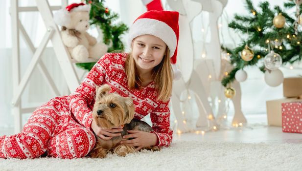 Child girl with dog sitting on floor with Christmas tree on background, looking at camera and smiling. Kid and pet doggy enjoying New Year time and decoration at home