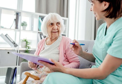 Elderly woman and nurse talking at home. Healthcare worker girl cares about senior female person indoors