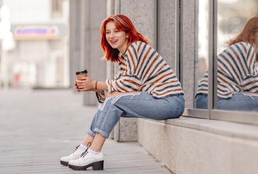Pretty girl with coffee cup sitting at street and smiling. Stylish trendy teenager outdoors with hot drink