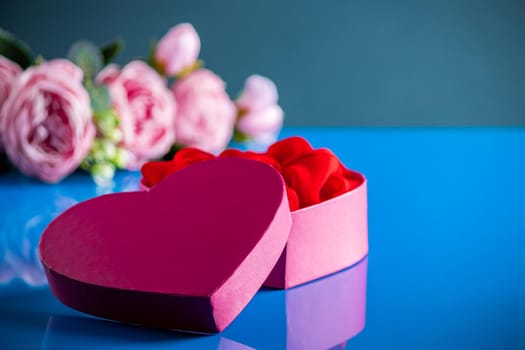 Heart shaped open gift box on blue background. Surprise for Valentine's Day. Open bright pink heart-shaped box and delicate flowers on a blue background.