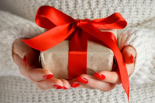 Gift with a red ribbon in hands on a white background. A woman in a white sweatshirt holds a holiday gift in her hands with a red ribbon.
