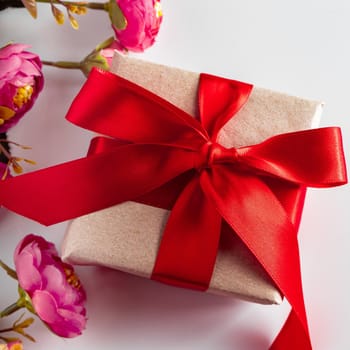 Gift box with red ribbon and flowers on a white background. Gift box with red ribbon and delicate pink flowers on a white background. Surprise on Women's Day and Mother's Day.