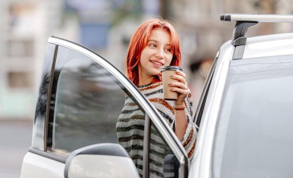 Pretty girl with coffee cup standing close to car at street and smiling. Stylish trendy teenager portrait outdoors