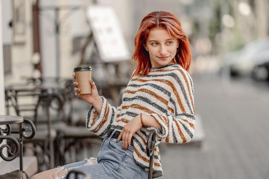 Pretty girl with coffee cup sitting in cafe and looking back at autumn. Stylish trendy teenager portrait with hot drink outdoors