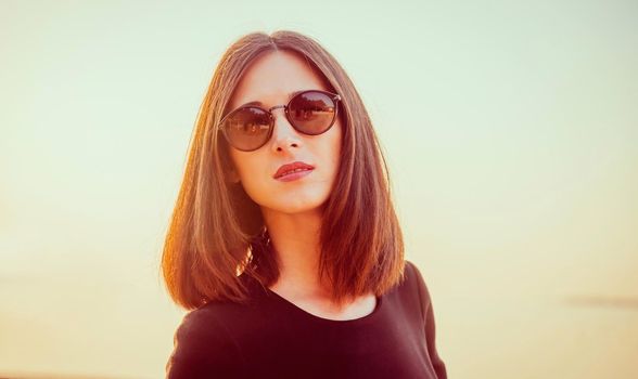 Portrait of attractive brunette woman with long hair in sunglasses on background of sea at sunny day. Image with sunlight effect