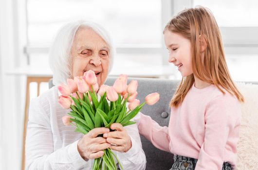 Grandmother sitiing on the sofa with preschool granddaughter, looking on flowers tulips bouquet and smiling