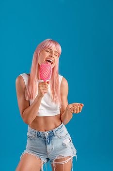 Funny young woman with pink hair holding hair comb and singing in it like a pop star while standing isolated over blue studio background. Beauty, hair care, lifestyle concept