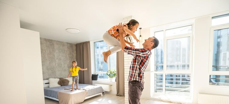 Overjoyed young father holding two little children siblings, having fun together at home, head shot. Excited dad playing with joyful adorable small daughter sisters, enjoying tender weekend moment.