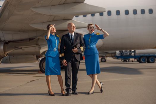 Full length shot of happy pilot standing together with two stewardesses in bright blue uniform in front of an airplane on a sunny day. Aircraft, aircrew, occupation concept