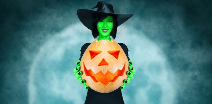 Smiling witch with green skin giving Halloween pumpkin on full moon background