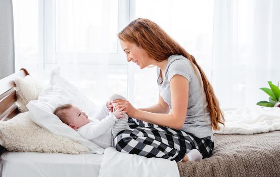 Beautiful young mother holding legs of her adorable little baby boy in her hands and looking at the child. Pretty girl with kid in the bed in morning time. Happy family moments