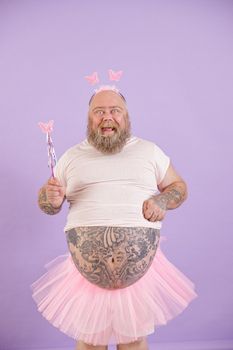 Funny emotional plus size man with bare tummy wearing fairy costume holds magic stick posing on purple background in studio