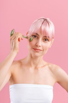 Pretty young woman with pink hair smiling at camera while using natural jade facial roller for skin care isolated over pink background. Face massage and beauty trends concept