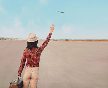 Traveler woman with suitcase waves her hand to airplane, rear view. Space for text in right part of image