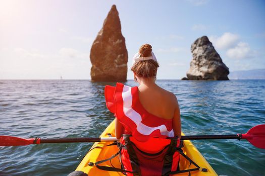 Young brunette woman in red swimsuit and Santa hat, swimming on kayak around basalt rocks like in Iceland. Back view. Christmas and travel concept