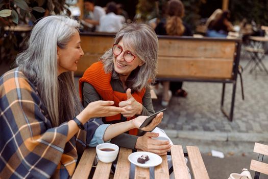 Smiling senior woman shows thumb up to friend holding cellphone at small table with coffee and candies in street cafe on nice autumn day