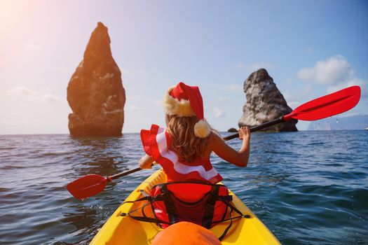Young brunette woman in red swimsuit and Santa hat, swimming on kayak around basalt rocks like in Iceland. Back view. Christmas and travel concept