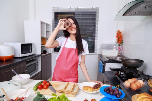 Cheerful woman is covering her eye with chocolated donut in kitchen room at home
