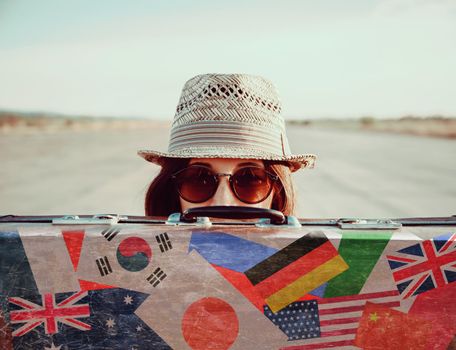 Hipster girl in a hat and sunglasses looks out from vintage suitcase. Suitcase with stamps flags of different country