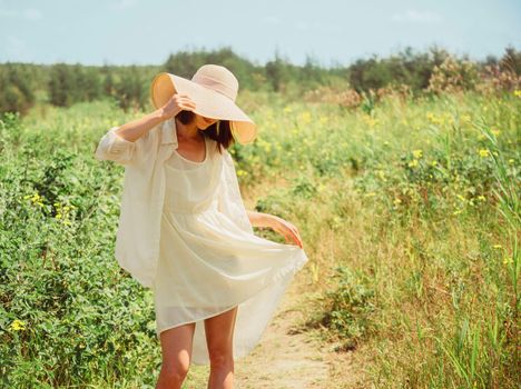 Happy beautiful young woman in yellow dress and hat with wide brim walking in summer park