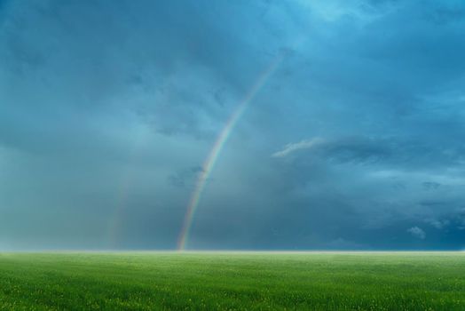 Rainbow in blue cloudy sky over green meadow in summer. Beautiful landscape
