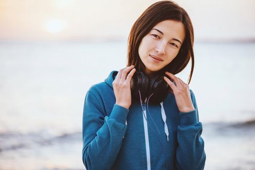 Beautiful young woman with headphones walking on coast near the sea at sunset