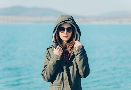 Beautiful young woman standing in a jacket with hood and sunglasses on the coast on background of sea