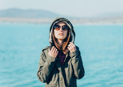 Beautiful young woman standing in a jacket with hood and sunglasses on background of sea
