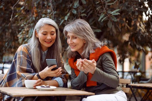 Asian lady shows photos on mobile phone to silver haired friend with cup of tea sitting at small table in street cafe