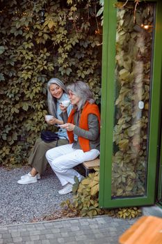 Pair of happy mature women friends with cups of drinks sits on bench near wall covered with green ivy. Long-time friendship