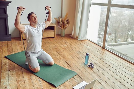 Top view of athletic male sitting on mat and exercising shoulders with resistance bands while watching notebook