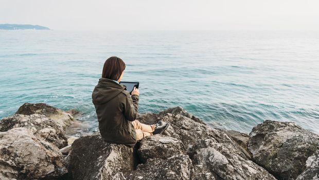 Traveler young woman sitting on coast near the sea and working on digital tablet