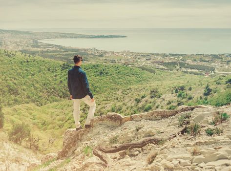 Young man standing on peak of mountain and looking at town around bay in summer, rear view