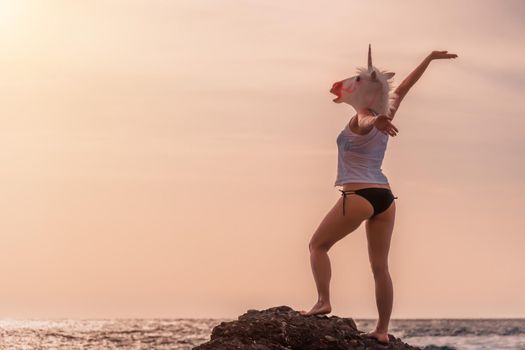 Unusual Woman with unicorn head mask dancing by the sea on the beach, strange funny weird photo.